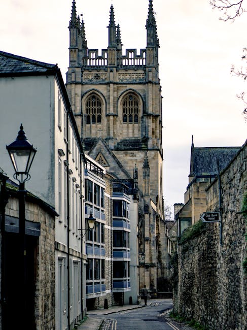 A historic stone building with large windows and a prominent tower featuring multiple spires, set against a partly cloudy sky with white fluffy clouds. In the foreground, there is a large evergreen tr