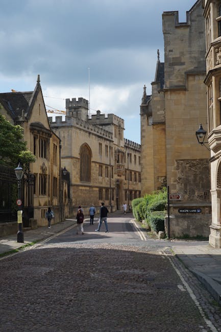 A cobbled street lined with historic stone buildings featuring gothic and medieval architectural elements, including crenellated towers and arched windows, under a partly cloudy sky. Several pedestria