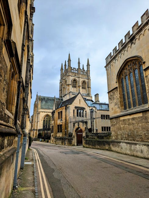 A cobbled street lined with historic stone buildings featuring gothic and medieval architectural elements, including crenellated towers and arched windows, under a partly cloudy sky. Several pedestria