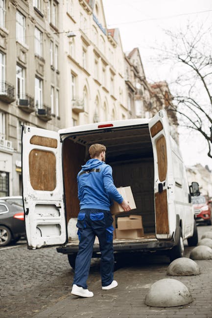 A man sits on the edge of an open white commercial van door, holding and reading a document. He is surrounded by several cardboard boxes, some stacked on the ground nearby and others inside the van, w