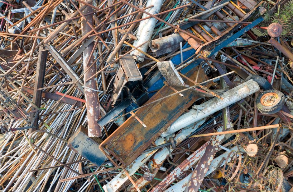 A large metal waste skip with a weathered surface, situated on a paved area next to a brick wall with some ivy growing over the top, in an outdoor urban setting. The skip is positioned for loading or