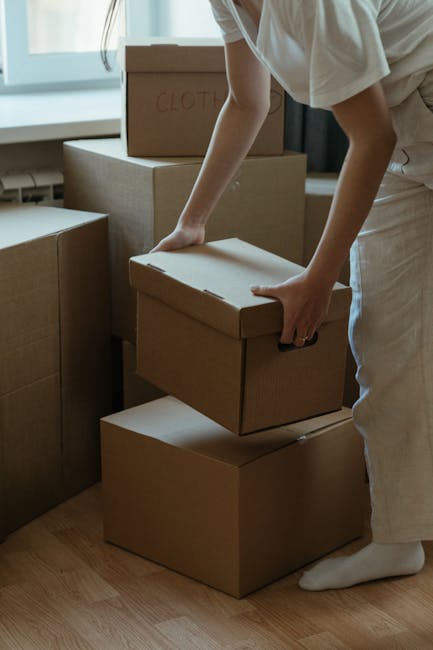 A close-up view of a person wearing a dark green shirt and dark blue trousers, securing a cardboard moving box with a red and white caution label that reads 'CAUTION THIS SIDE UP' with an upward point
